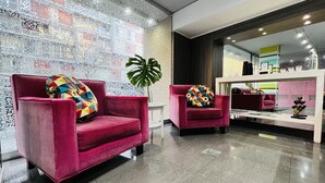 Eine moderne Lobby mit zwei bequemen roten Sesseln, jeder mit einem farbenfrohen geometrischen Kissen. Eine große grüne Pflanze bringt einen Hauch von Natur herein. Der Boden ist mit poliertem Fliesenbelag versehen, und die Wände zeigen ein dekoratives geometrisches Muster.