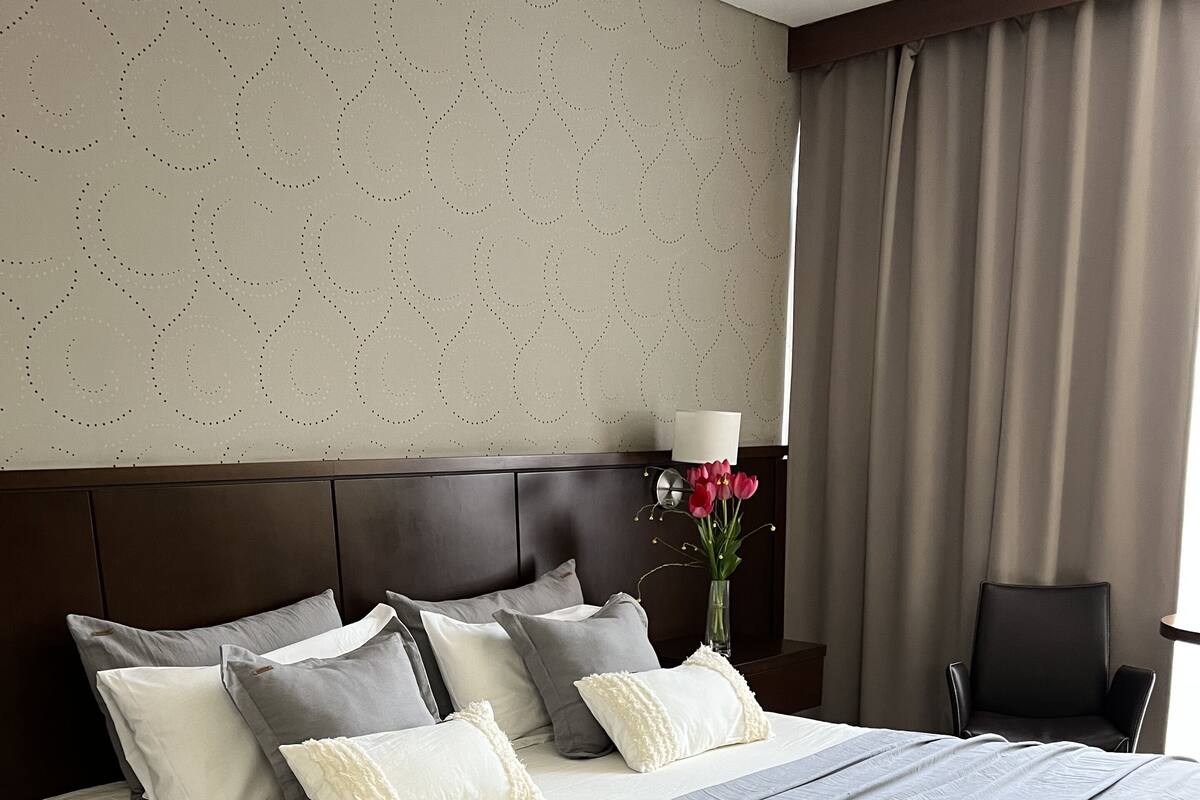 Una cama bien hecha con ropa de cama gris y blanca, un cabecero oscuro y un florero con flores sobre la mesita de noche.