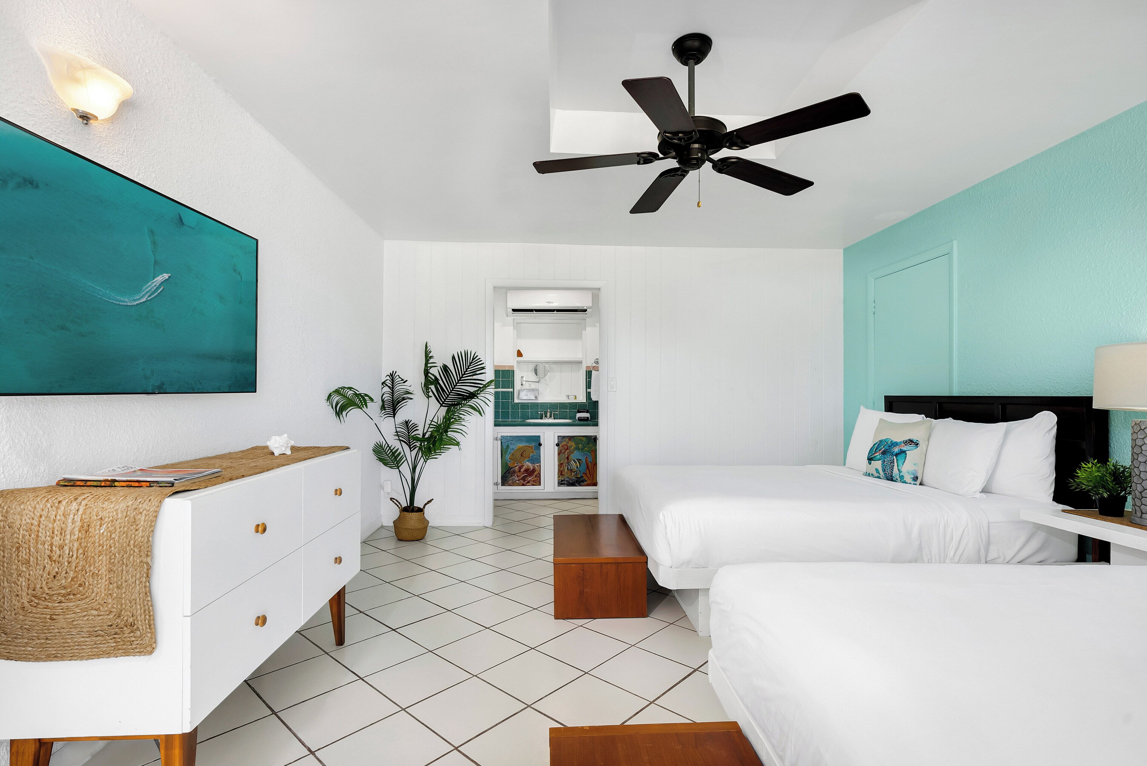Un dormitorio moderno con dos camas, un ventilador de techo, un televisor y una kitchenette al fondo.