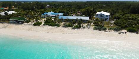 An aerial view of a beachfront property with multiple buildings, including a large house with a blue roof and a smaller structure with a green roof, surrounded by lush greenery and a sandy beach.