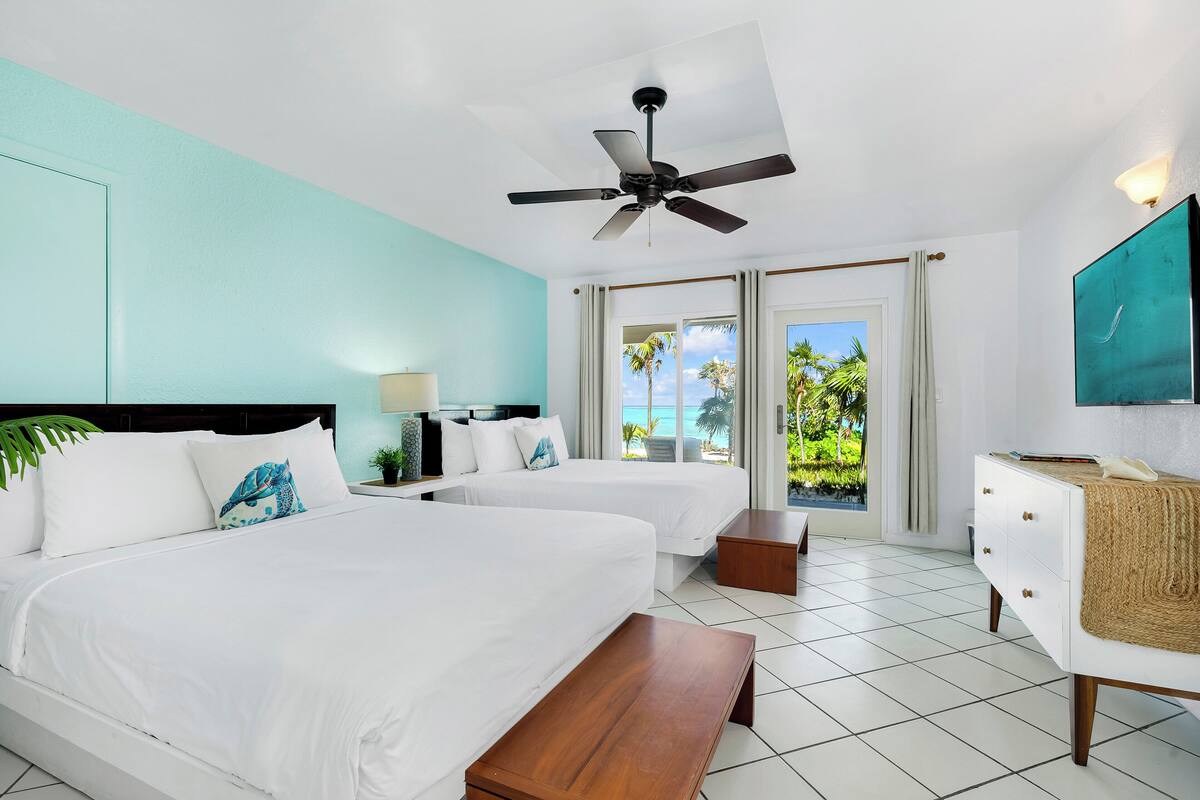 A hotel room with two beds, a ceiling fan, a view of the ocean, and a TV on the wall.