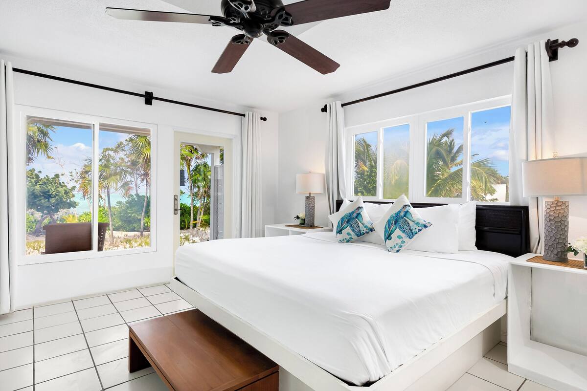 A bedroom with a large bed, a ceiling fan, and a view of palm trees through the windows.