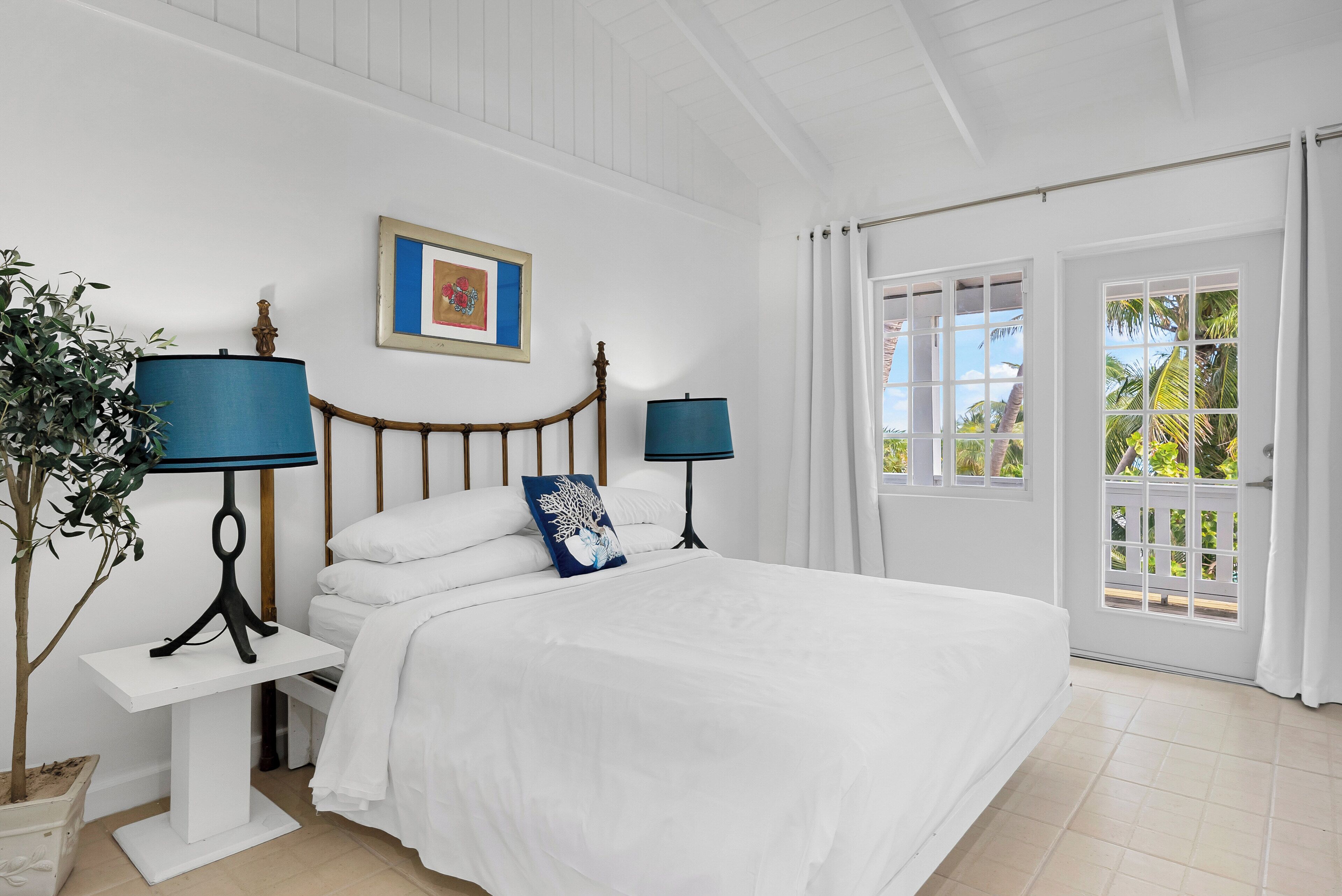 A bedroom with a bed, two bedside tables with lamps, a potted plant, a framed picture on the wall, and a view of the outdoors through a door.