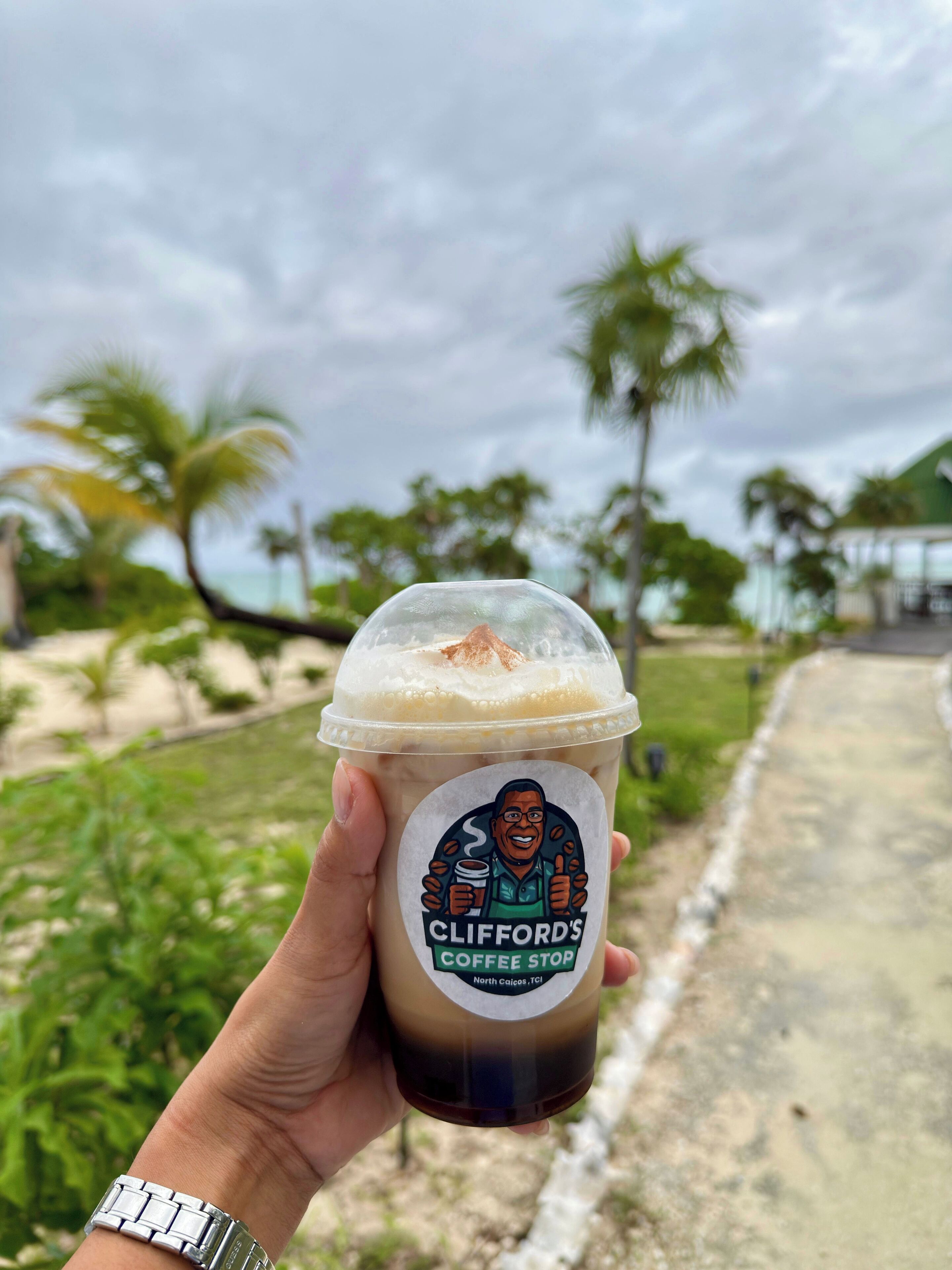 Une main tenant une tasse de café glacé avec une étiquette indiquant « Clifford’s Coffee Stop ».
