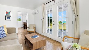 A spacious living room with a sofa, a wooden coffee table, and a chair. There is a large window with curtains, a door leading outside, and a small table with a decorative item.