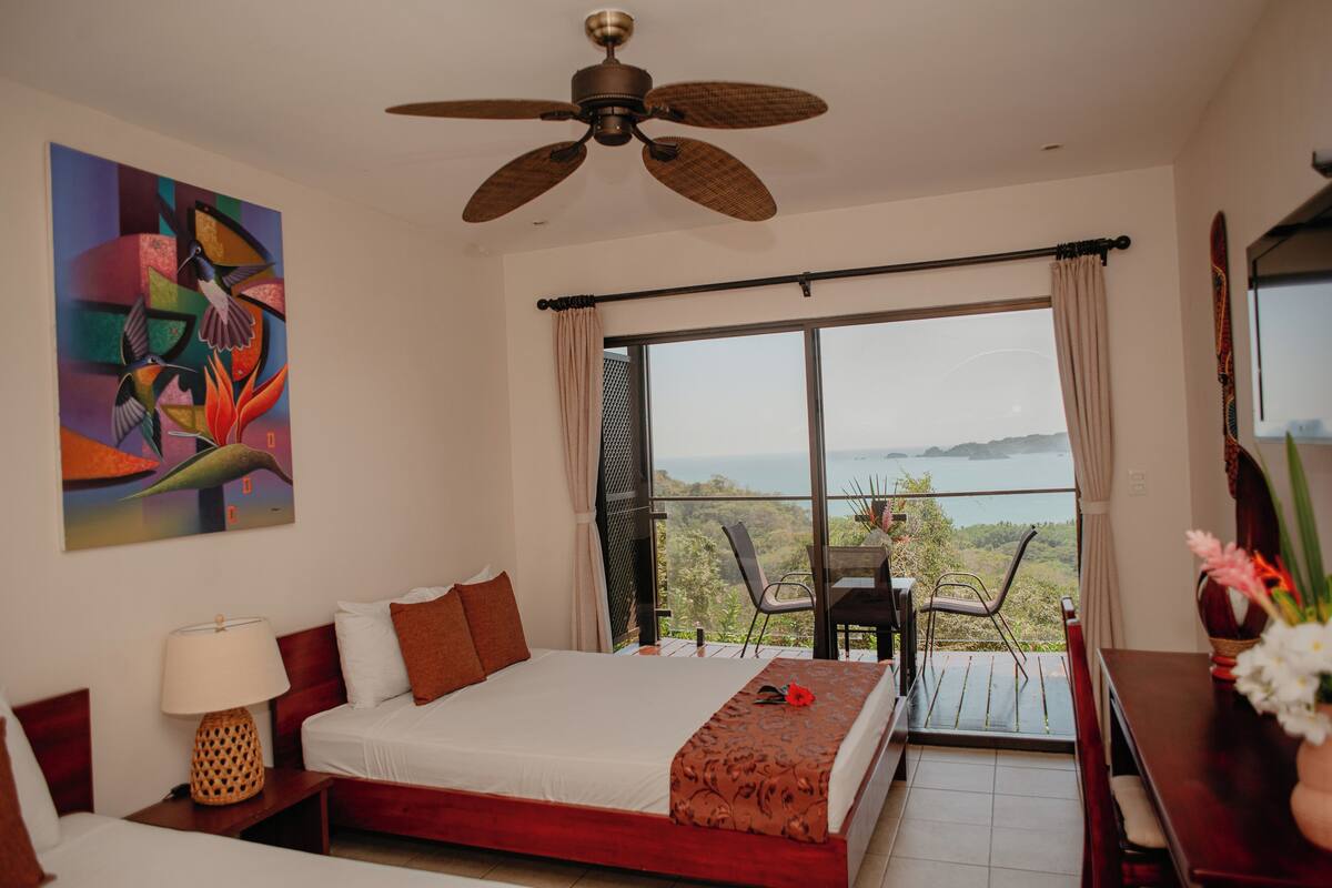 Un dormitorio con cama, ventilador de techo, ventana con vistas y un cuadro en la pared.