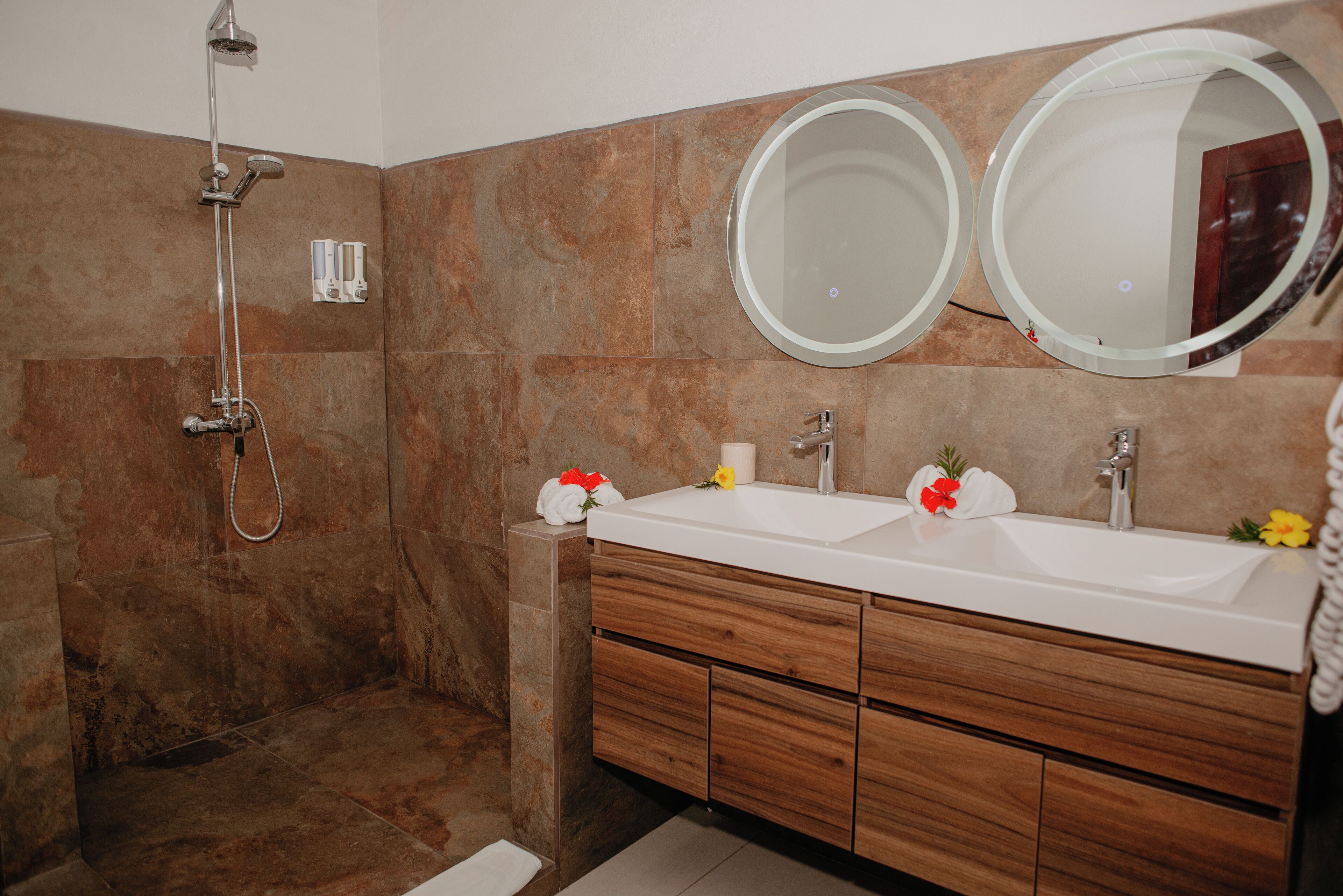 A bathroom with a shower, two sinks, wooden cabinets, and two round mirrors.
