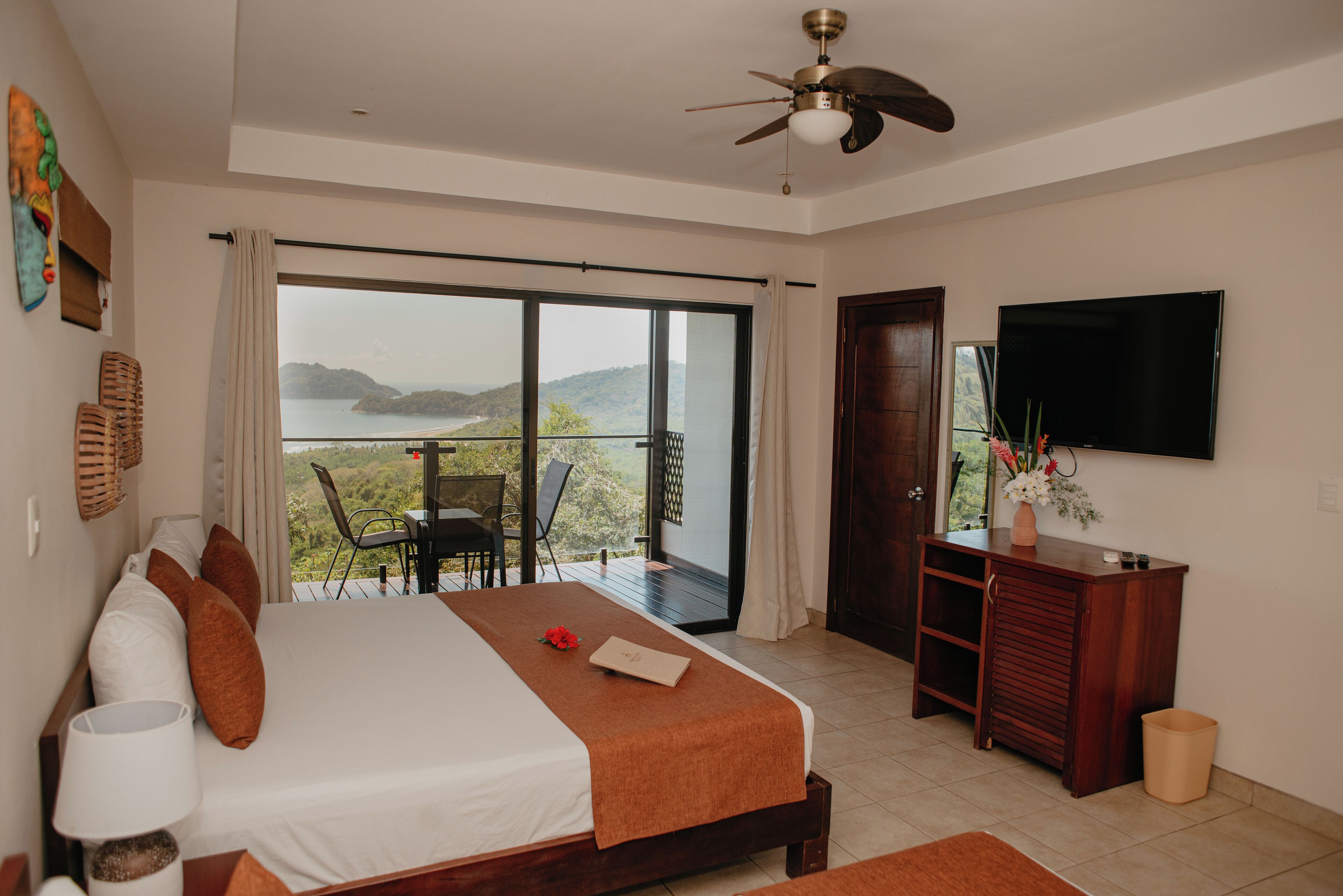 A bedroom with a large bed, a ceiling fan, a TV, and a balcony with a view.