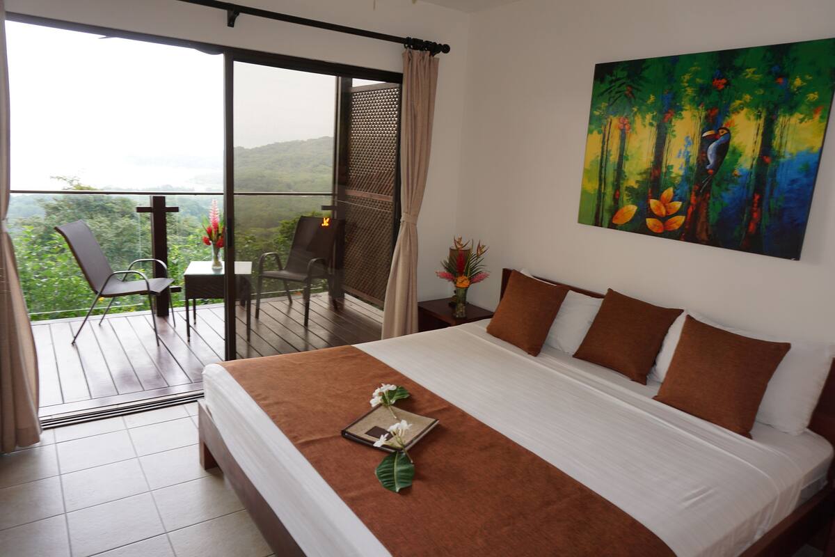 Un dormitorio con una cama grande, un cuadro, un balcón con sillas y vistas al exterior.