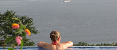 A person in a swimming pool overlooking a calm sea with a sailboat in the distance.