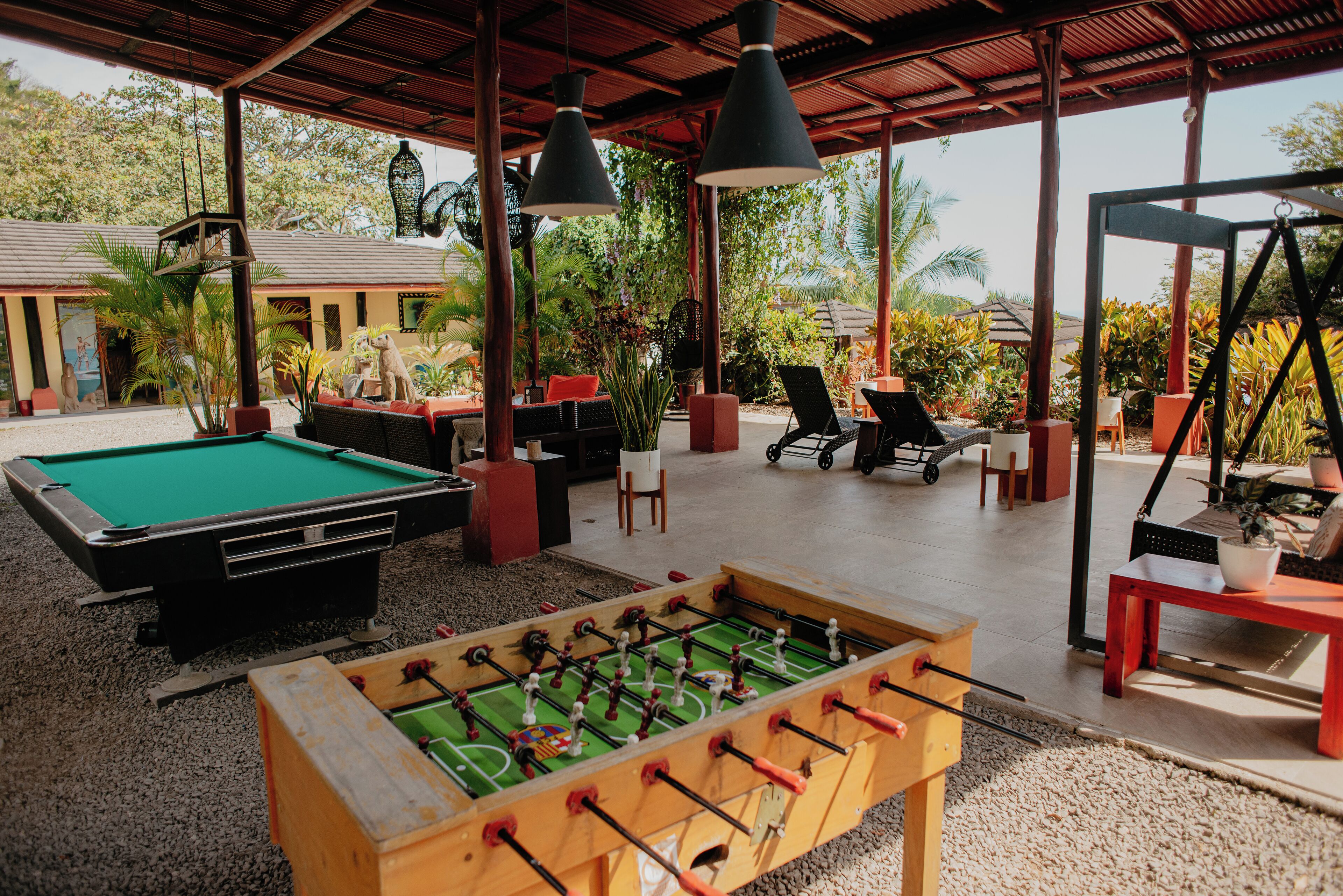 Una mesa de billar, una mesa de fútbol de mesa y zona de estar al aire libre bajo una zona cubierta, con plantas y árboles de fondo.