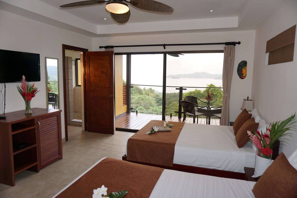 Habitación de hotel con dos camas, televisión, escritorio y balcón con vistas.