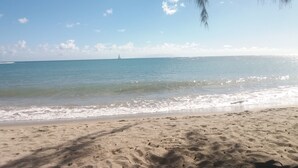 Una playa con cielos despejados, agua tranquila y un velero a lo lejos.