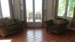 Un salón amplio con suelo de baldosas, dos sillones verdes, un sofá de mimbre y un ventanal corredero que da a una zona exterior con sillas.