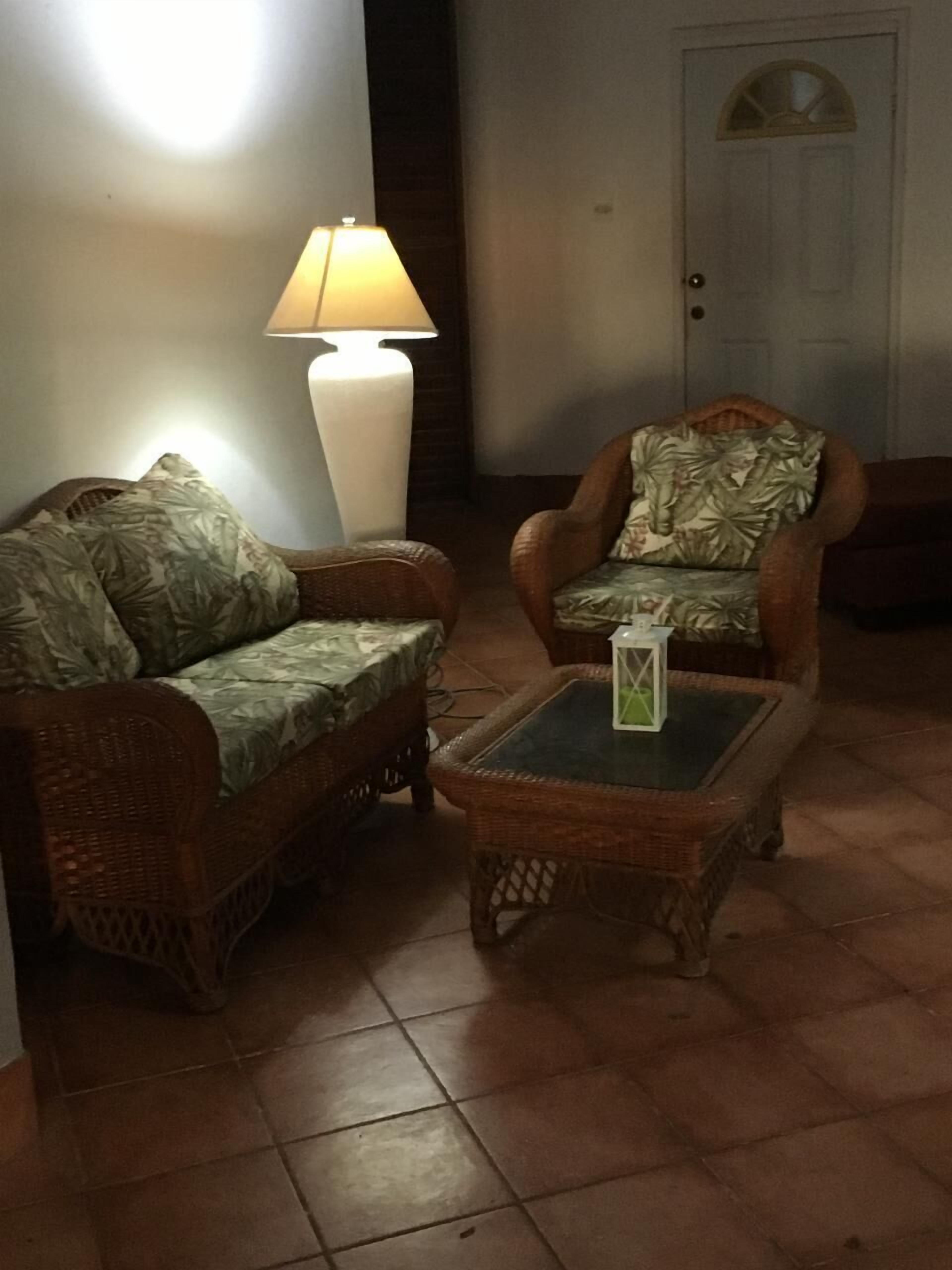 Een kamer met twee rieten banken, waarvan één met een bloemenkussen en de andere met een bladmotief, een salontafel met glazen blad, en een hoge vloerlamp.