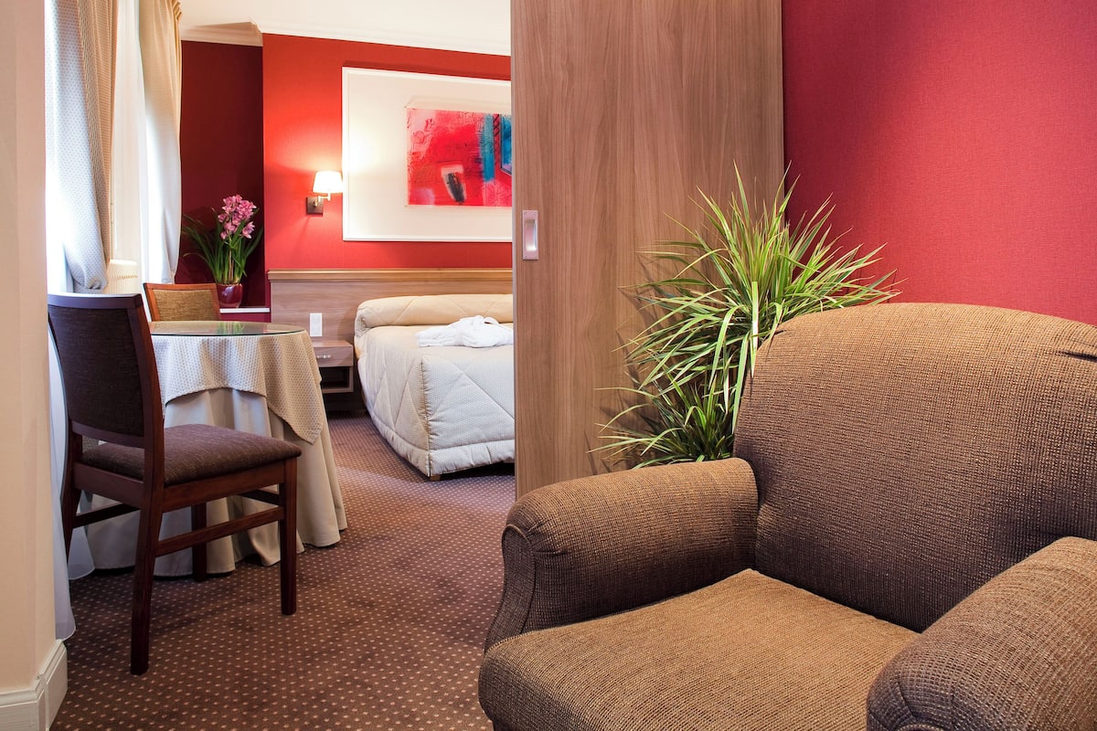 Habitación de hotel con sofá marrón, una mesita con una silla, cama con edredón blanco, planta en maceta y una pared de acento roja.