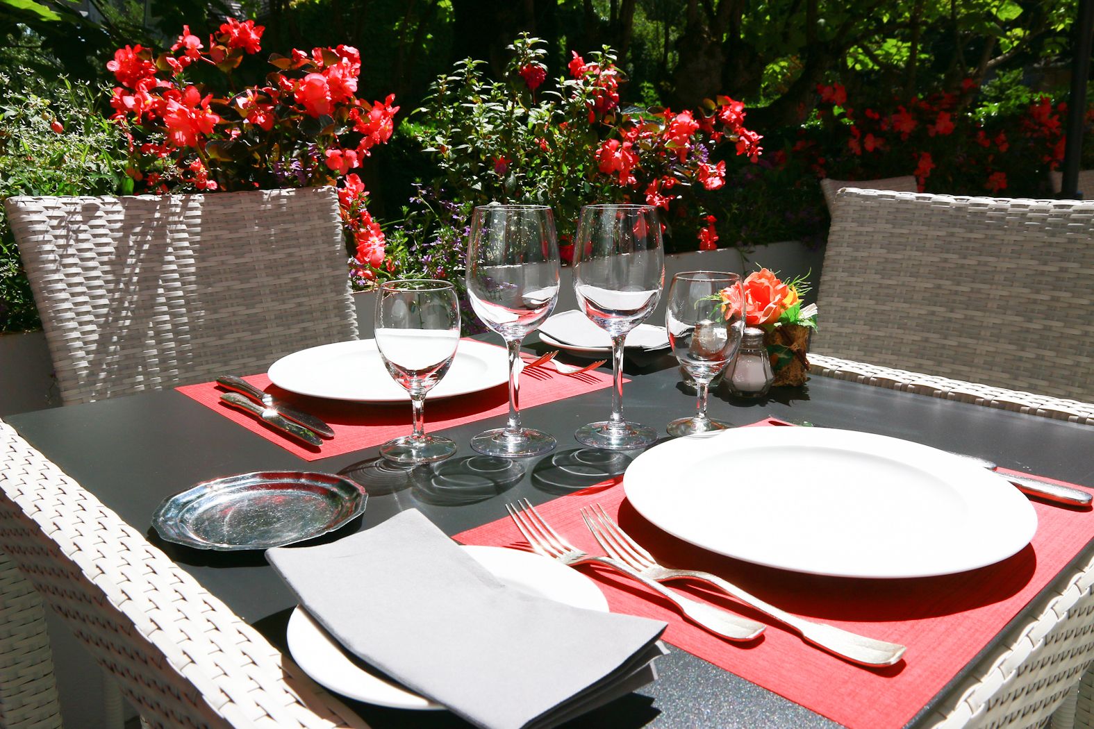 Une table à manger d’extérieur, dressée avec de l’assiette blanche, des verres à vin transparents et des serviettes rouges.