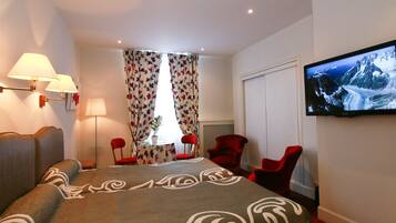 Une chambre d’hôtel avec un lit, deux fauteuils rouges, une télévision et une fenêtre avec des rideaux.