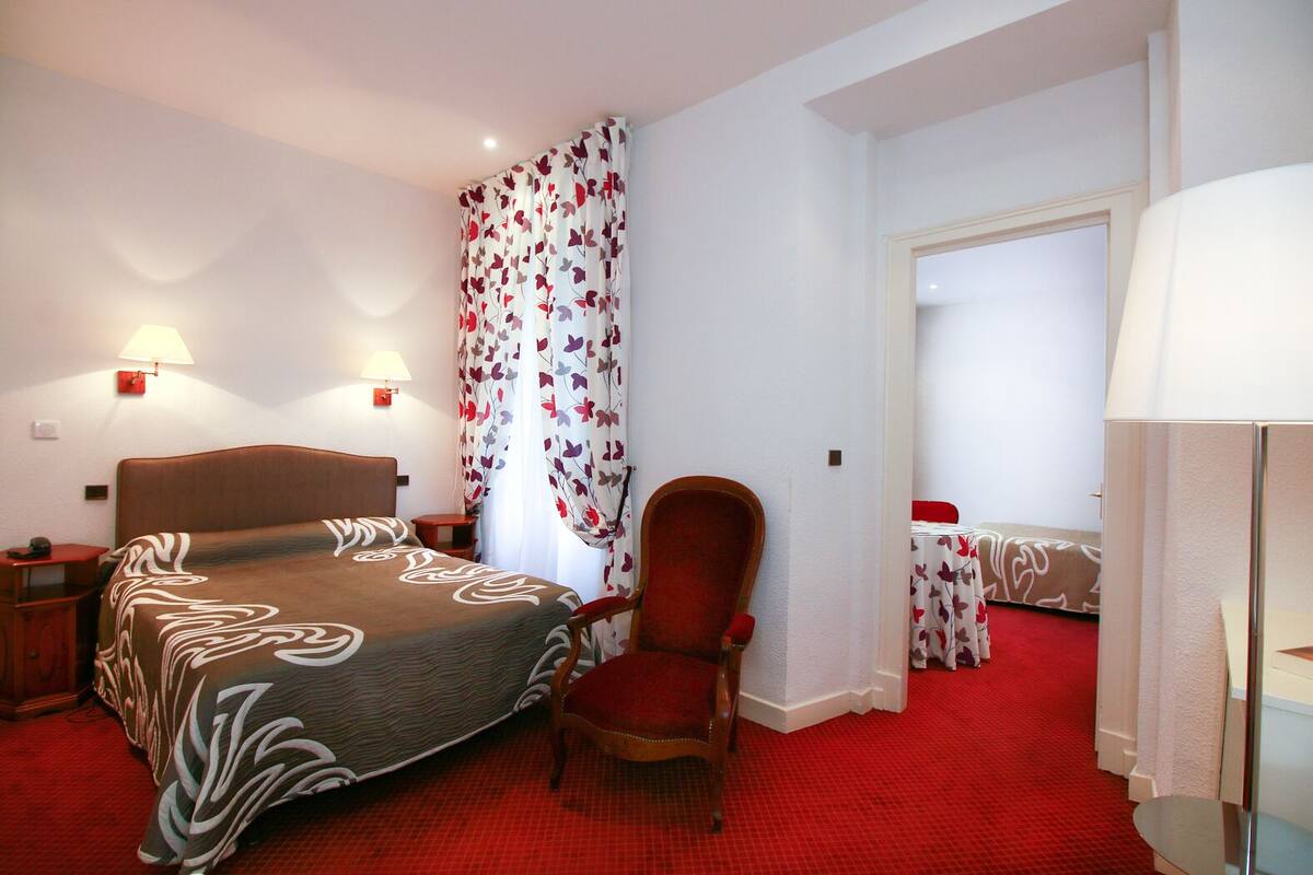 Una habitación de hotel con una cama, una silla, una mesita de noche, una lámpara y una puerta que da a otra habitación.