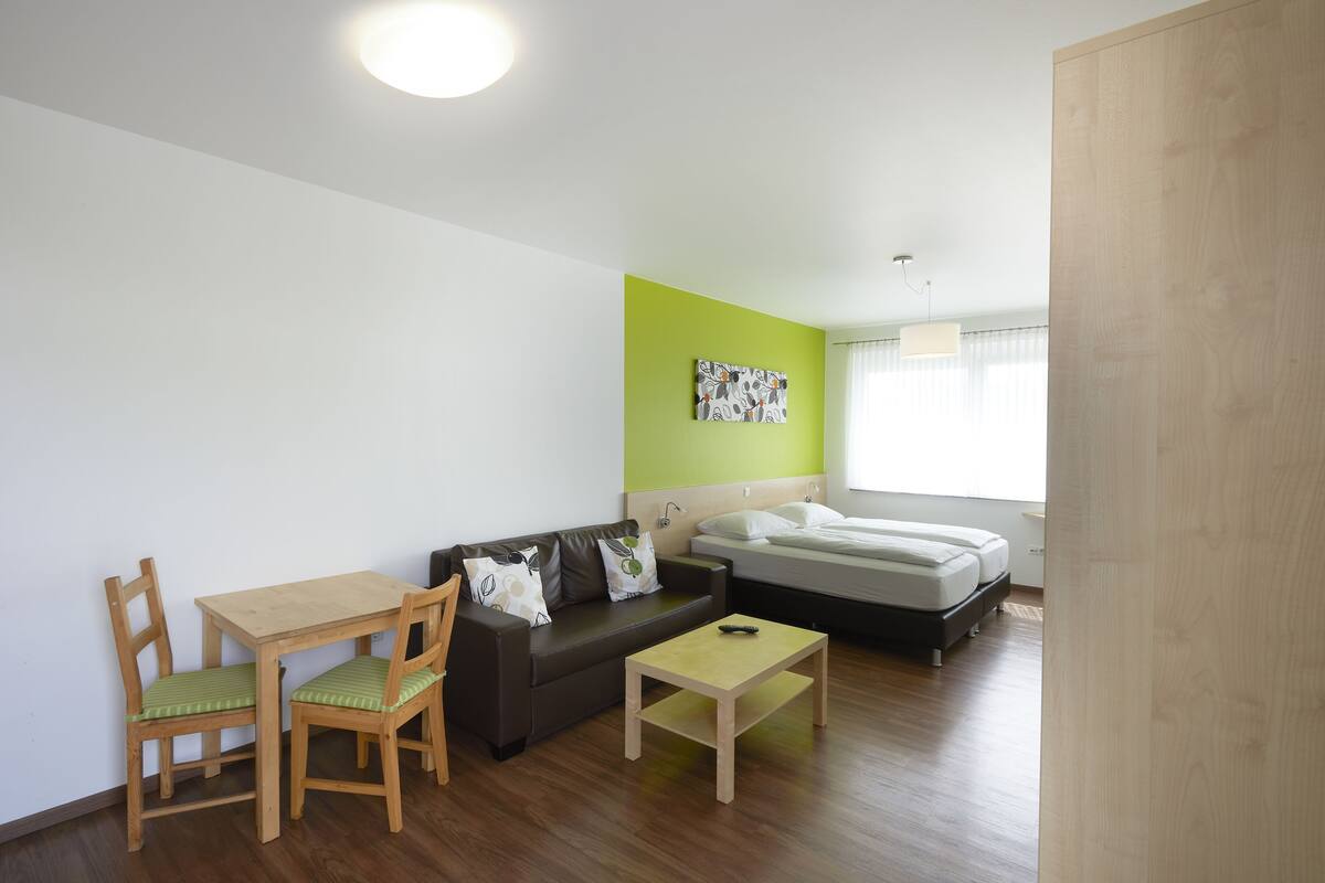 A small, modern room with a wooden floor, a green accent wall, a bed, a sofa, a table, and two chairs.