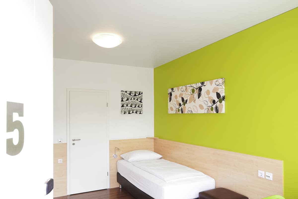 A modern hotel room with a wooden floor, a bed, a green chair, and a wall with a floral pattern.