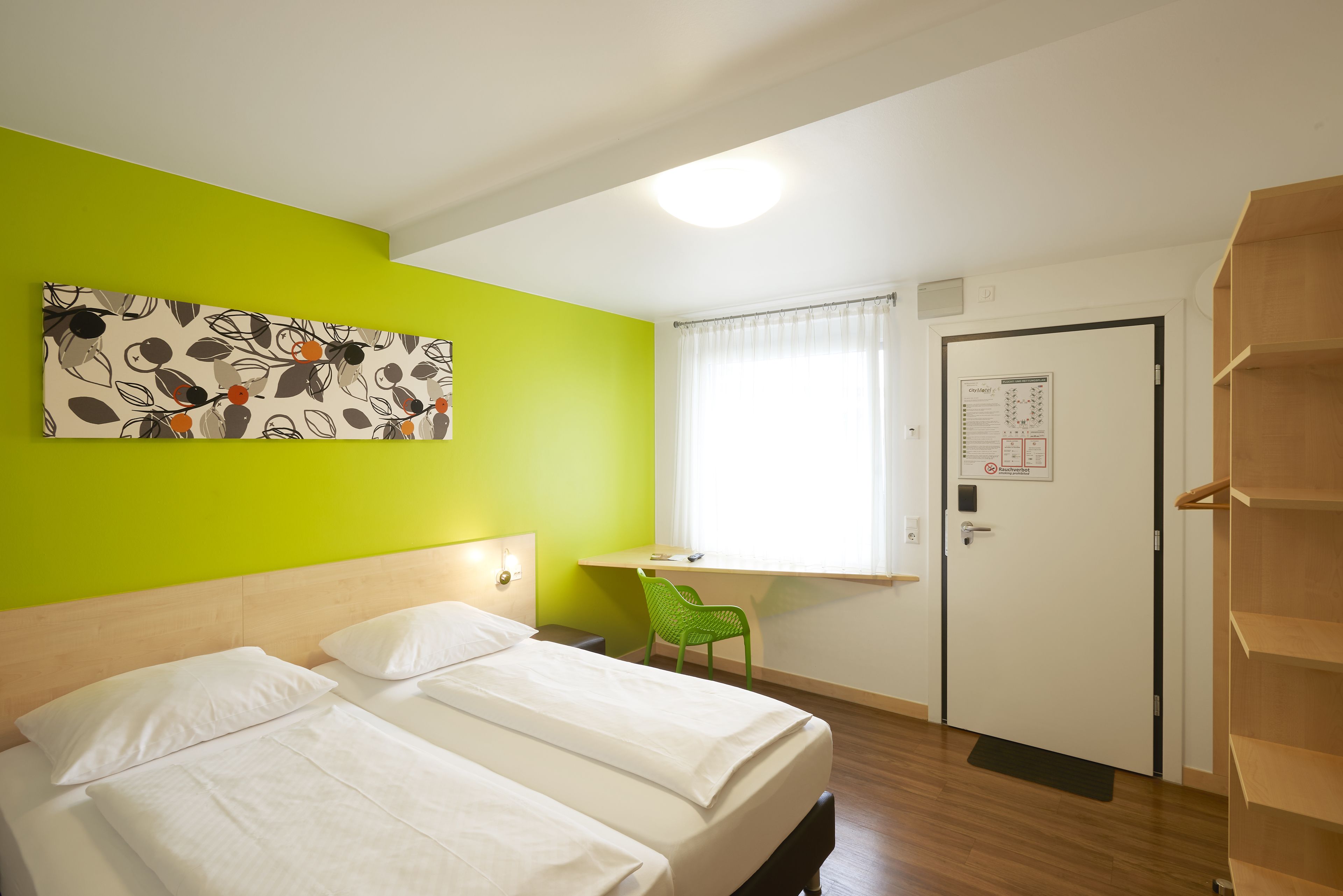A hotel room with two beds, a desk, a chair, a door with a notice, and a bright green wall.