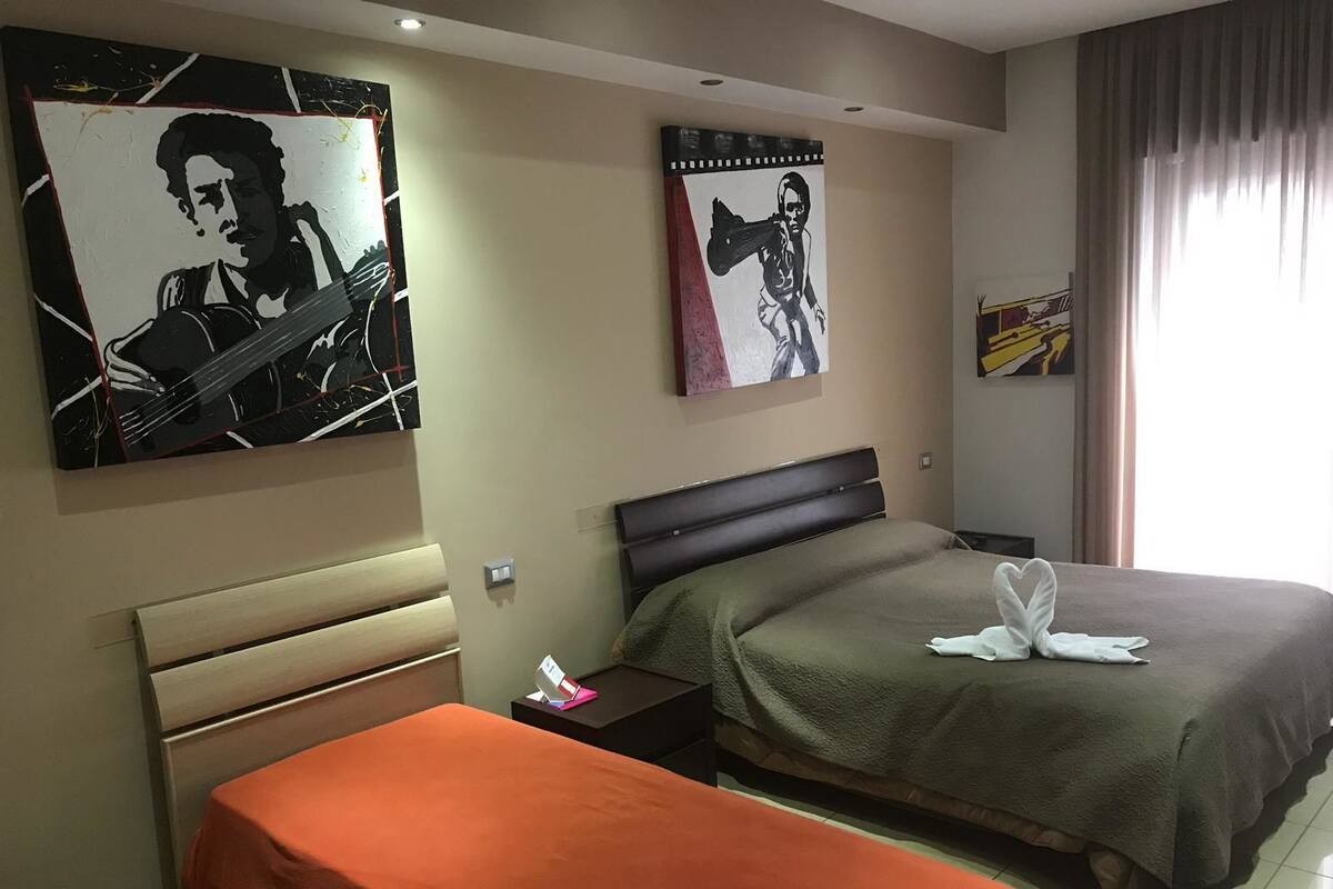 A hotel room with two beds, one with a green blanket and the other with an orange blanket. There are two paintings on the wall, one depicting a person playing a guitar and the other a person with a guitar.