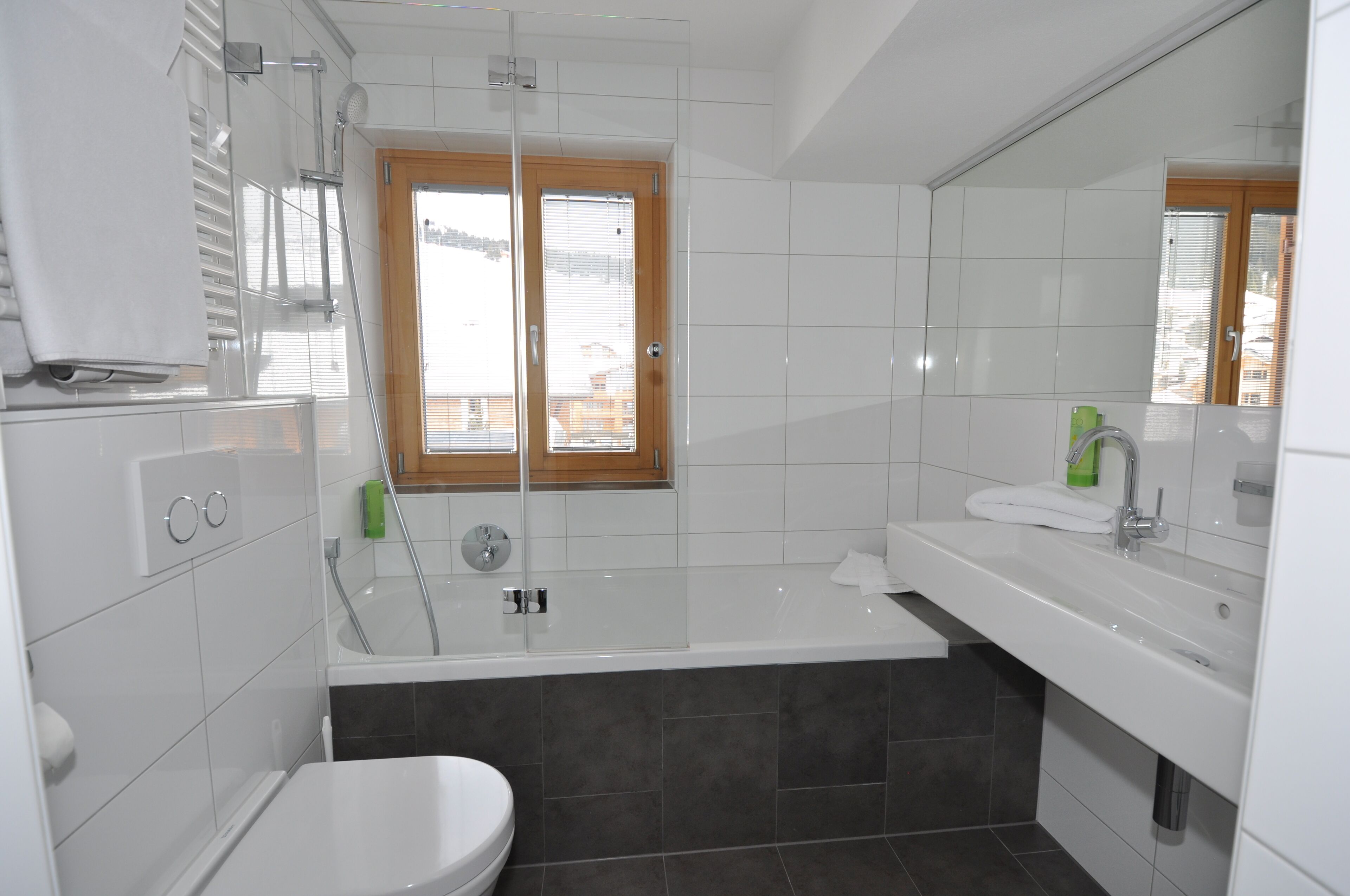 Un baño moderno con bañera, ducha y retrete. Cuenta con una ventana con persianas, un espejo y un perchero para toallas.