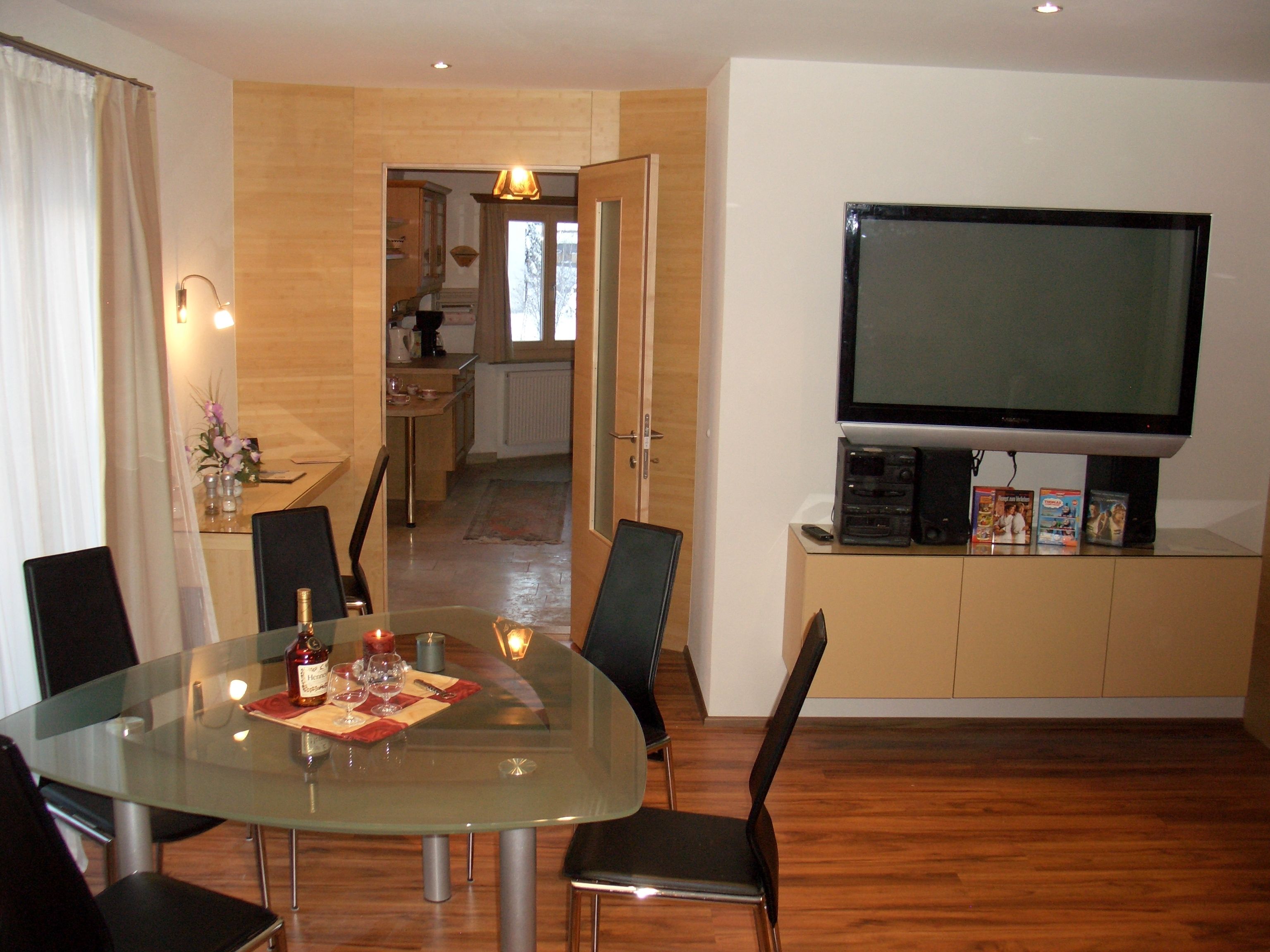 Un comedor con mesa de vidrio, sillas negras y un televisor sobre un mueble.