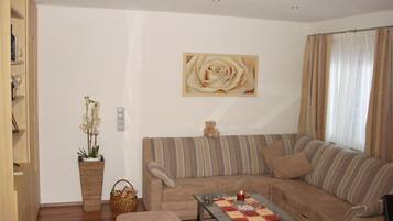 Uma sala de estar com um sofá, uma mesa de centro de vidro, uma mesinha com uma vela, um ursinho de pelúcia e um quadro de uma rosa pendurado na parede.