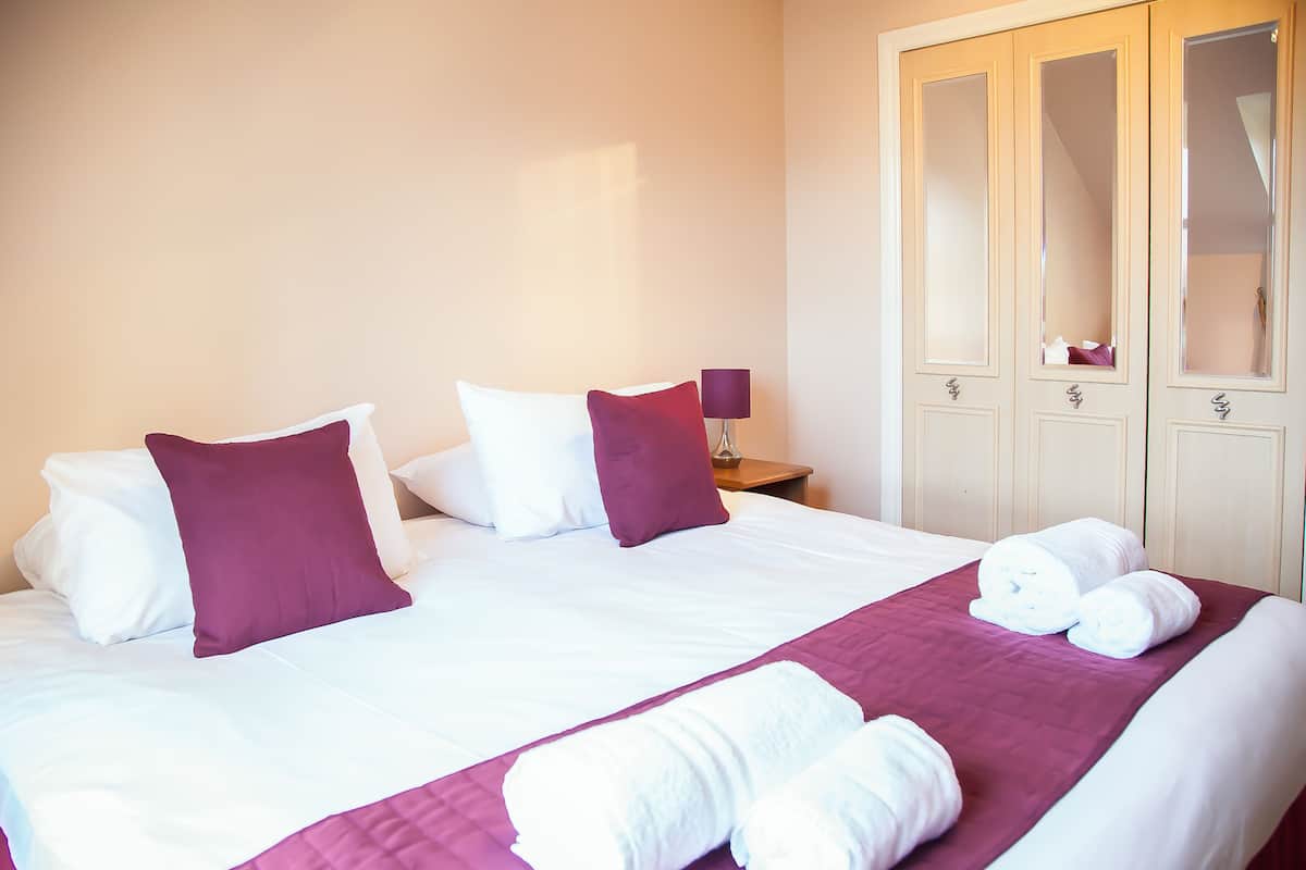 Ein Hotelzimmer mit einem großen Bett, Bettwäsche in Weiß und Lila, einem Nachttisch aus Holz und einem Schrank mit Spiegeln.