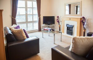 A living room with a sofa, a glass coffee table, a flat-screen TV, a fireplace, and a large window with curtains.