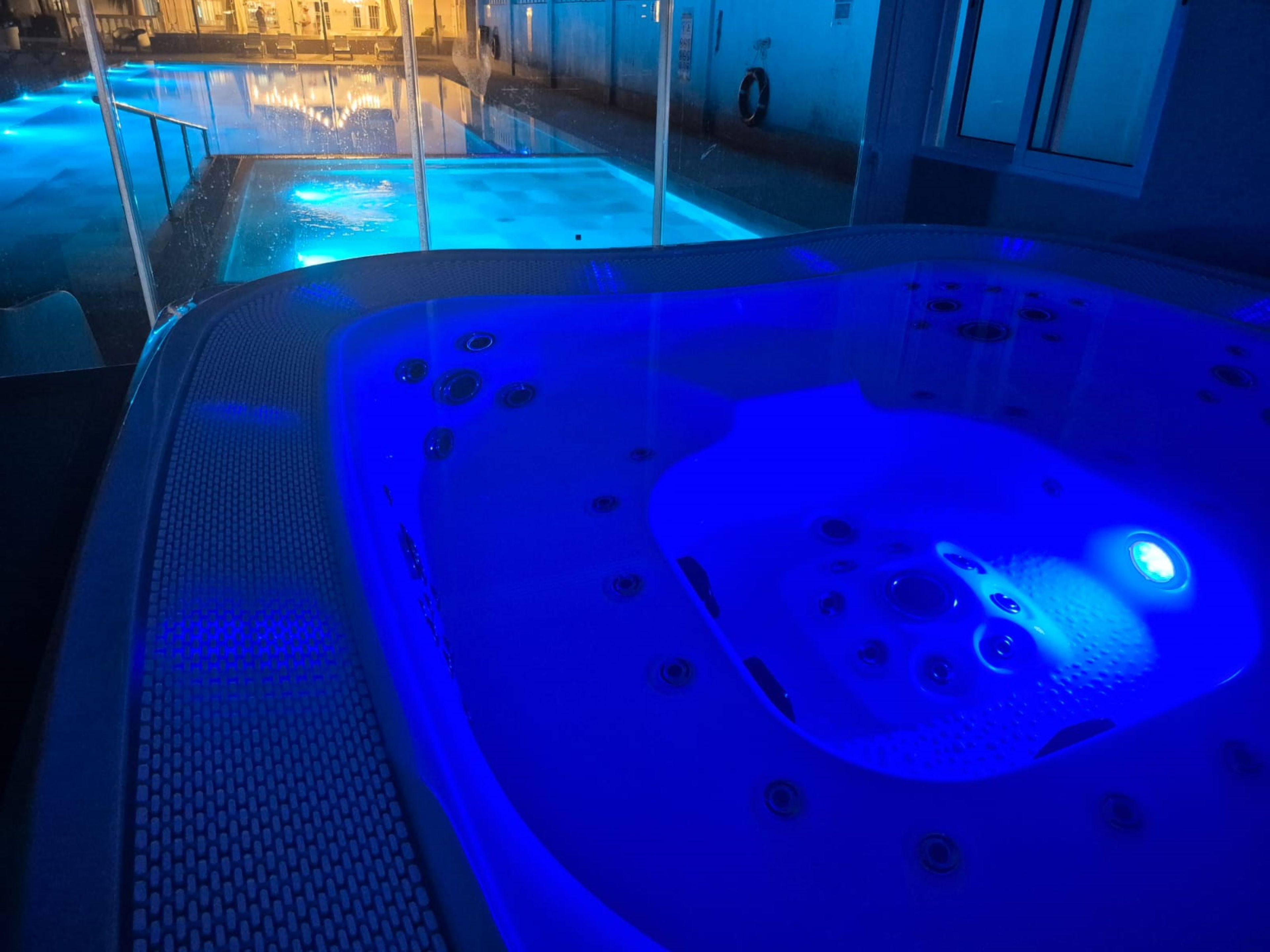 A hot tub with a blue light, a glass enclosure, and a view of a pool area.