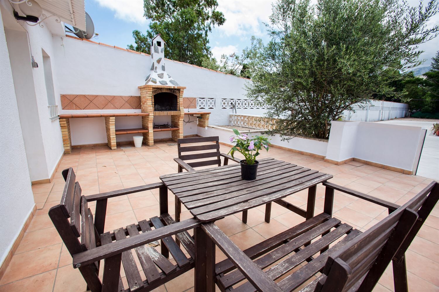 An outdoor patio with a stone barbecue, wooden table and chairs, and a potted plant.