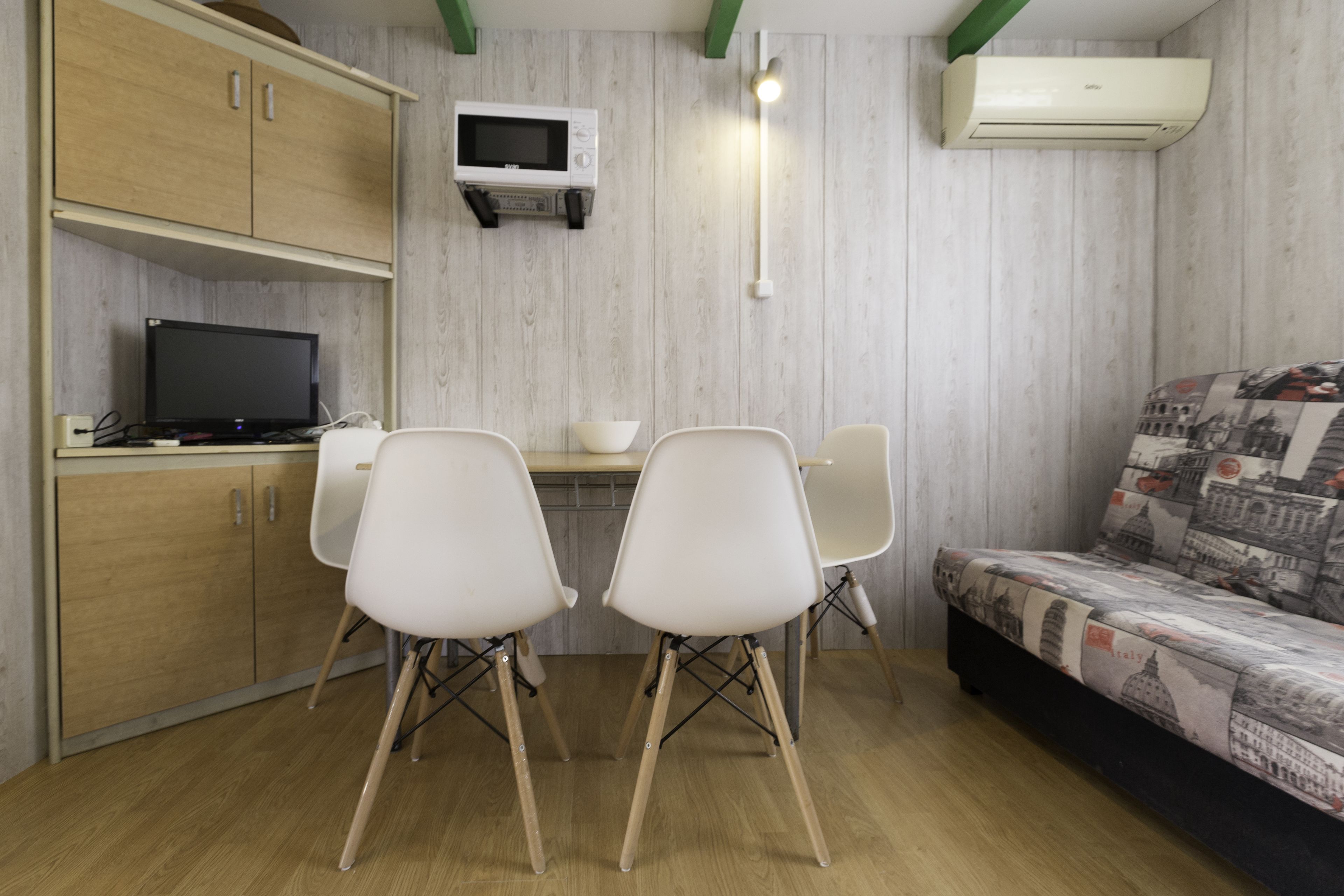Une pièce comprenant un meuble en bois, un téléviseur, un bureau avec deux chaises et un canapé recouvert d’un tissu à motifs.
