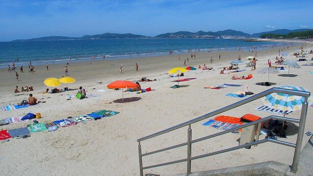 A beach with umbrellas, people sunbathing, and a staircase leading down to the beach. - Camping Los Llanos (Denia)