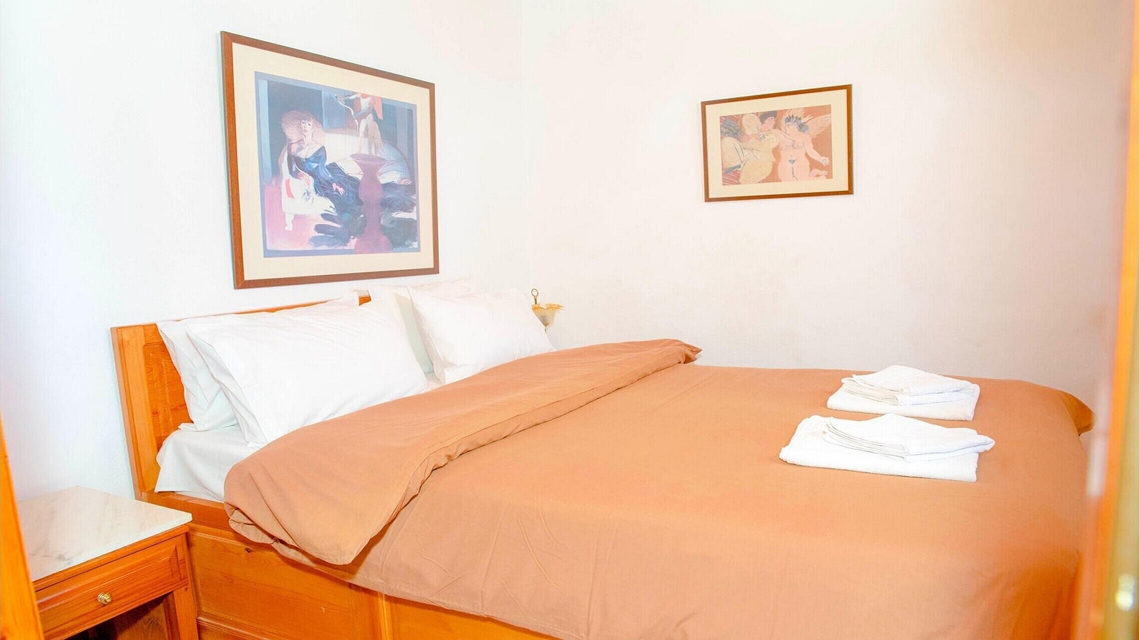 Un letto rifatto con un copriletto marrone, cuscini bianchi e due asciugamani piegati sul lato. Sopra il letto, due quadri appesi al muro.