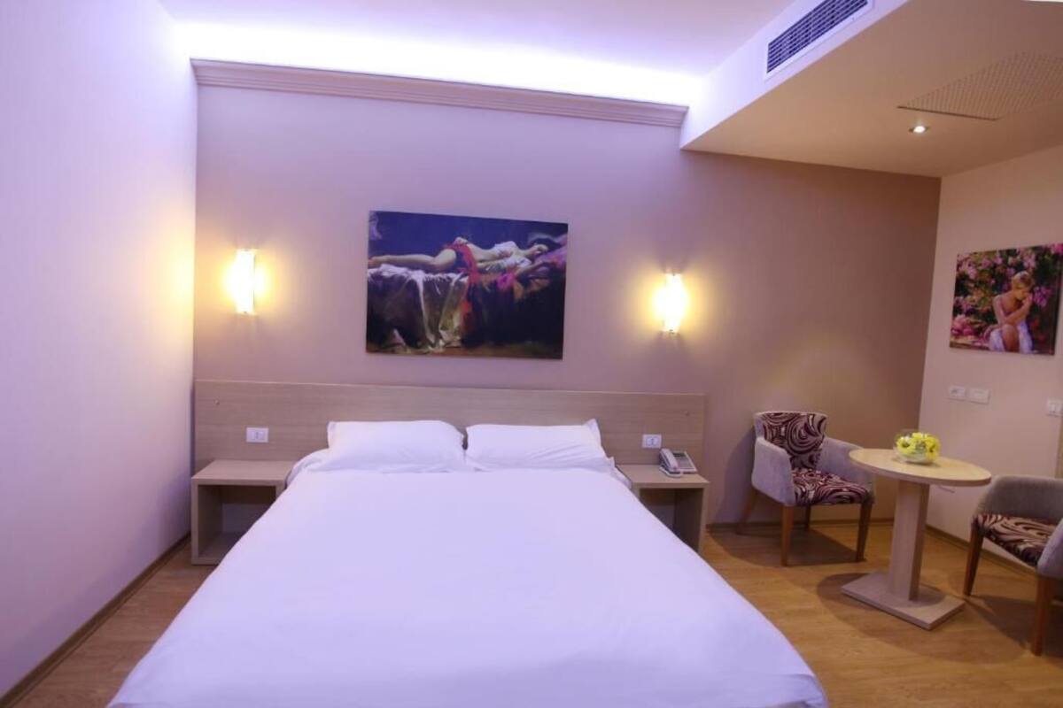 A hotel room with a large bed, two bedside tables, a chair, a small table, and two wall-mounted lamps.