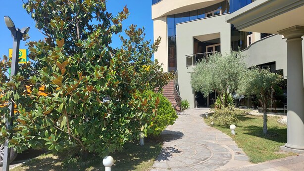 A pathway leading to a modern building with a glass facade and a landscaped garden area with trees and shrubs. - Hotel Green (Vaqarr)