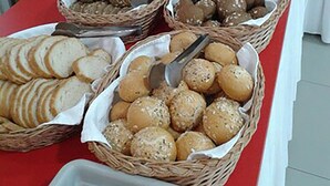 A buffet table with various breads, rolls, and pastries in baskets, along with a container of utensils and a pitcher of orange juice. - Hotel Green (Vaqarr)