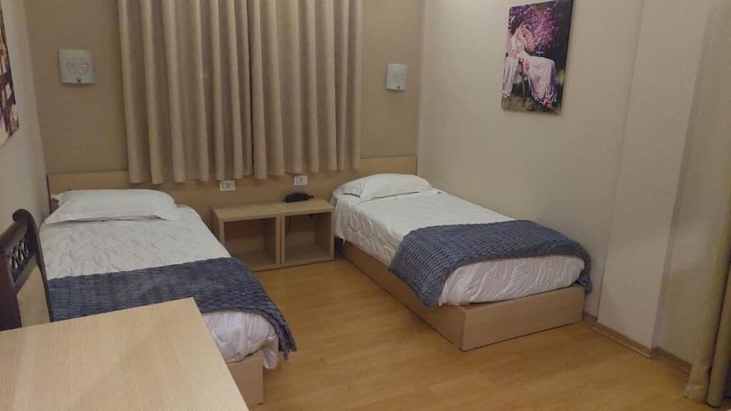 Quarto pequeno e limpo com duas camas de solteiro, uma mesa de madeira e uma escrivaninha com cadeira.