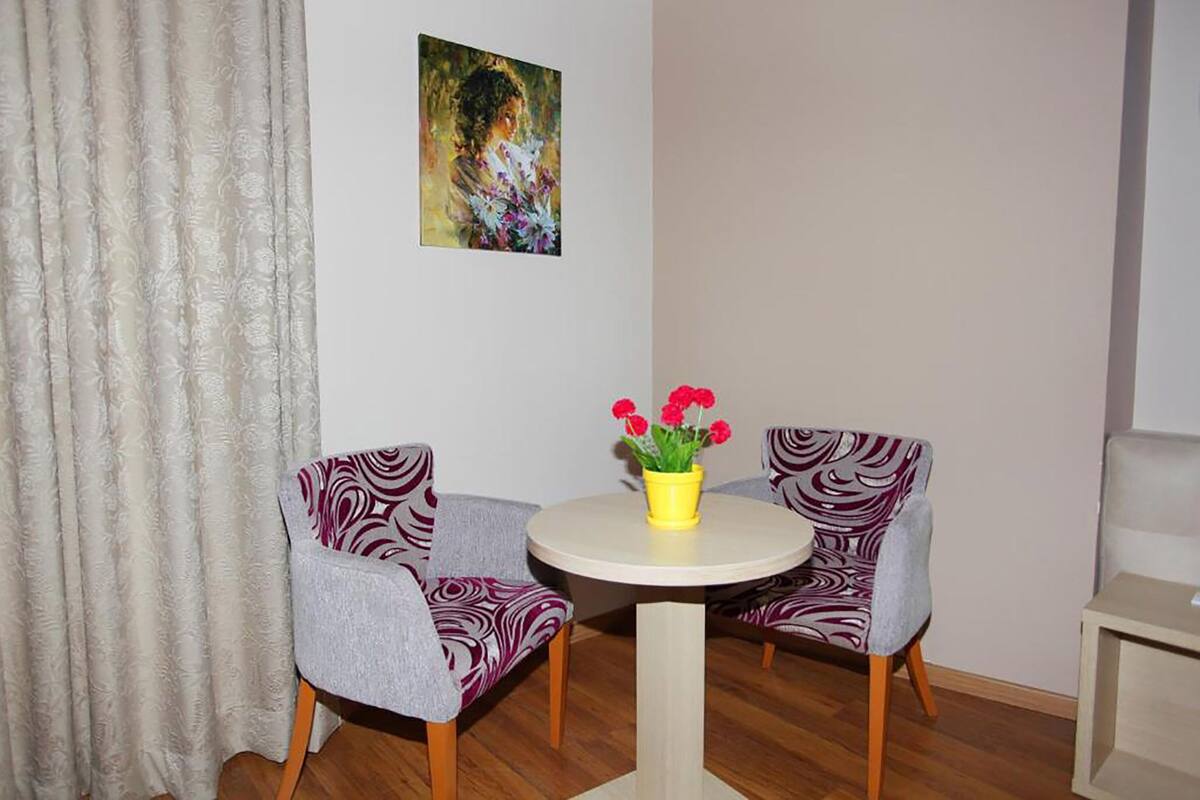 A room with two patterned armchairs, a round table with a yellow vase of flowers, and a painting on the wall.