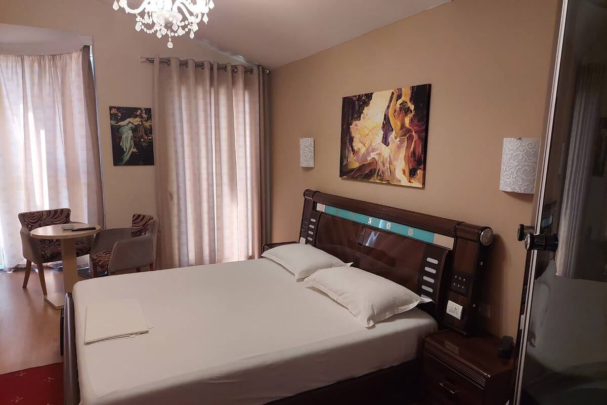 A hotel room with a large bed, a bedside table, a chair, a small table, and a painting on the wall.