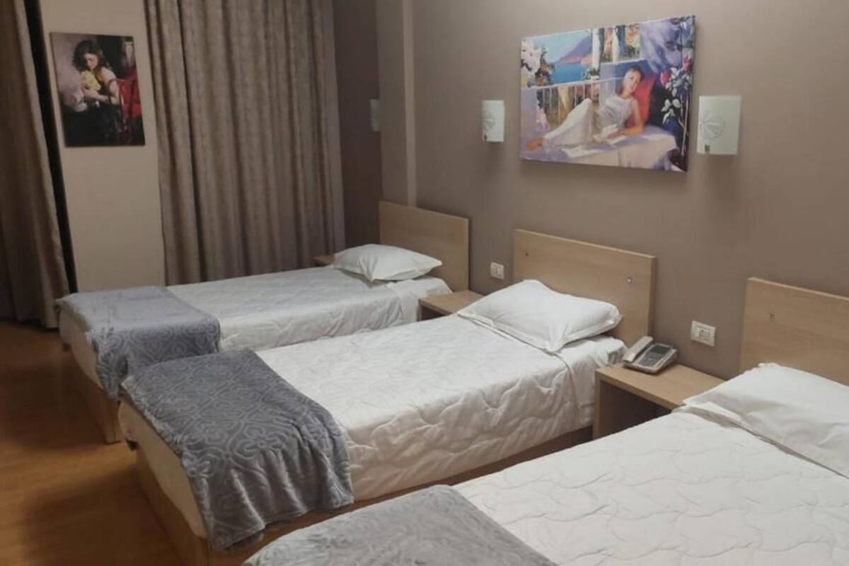 A hotel room with three single beds, each with a pillow and a blanket. There is a bedside table with a phone on it. The room has a neutral color scheme with a painting on the wall.