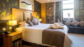 Una habitación de hotel con una cama grande, una mesita de noche con lámpara, una estantería, un sofá y una ventana con cortinas.