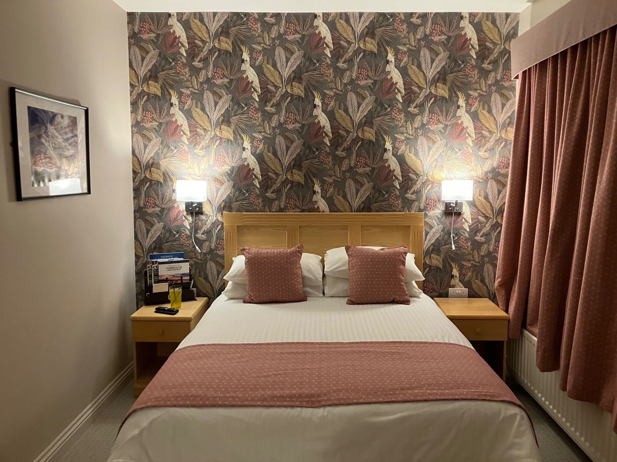 Habitación de hotel con una cama grande, dos mesitas de noche, un buró, un cuadro en la pared y un papel tapiz floral.
