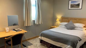 Habitación de hotel con cama, escritorio, silla, lámpara, un cuadro en la pared y ventana con cortinas.
