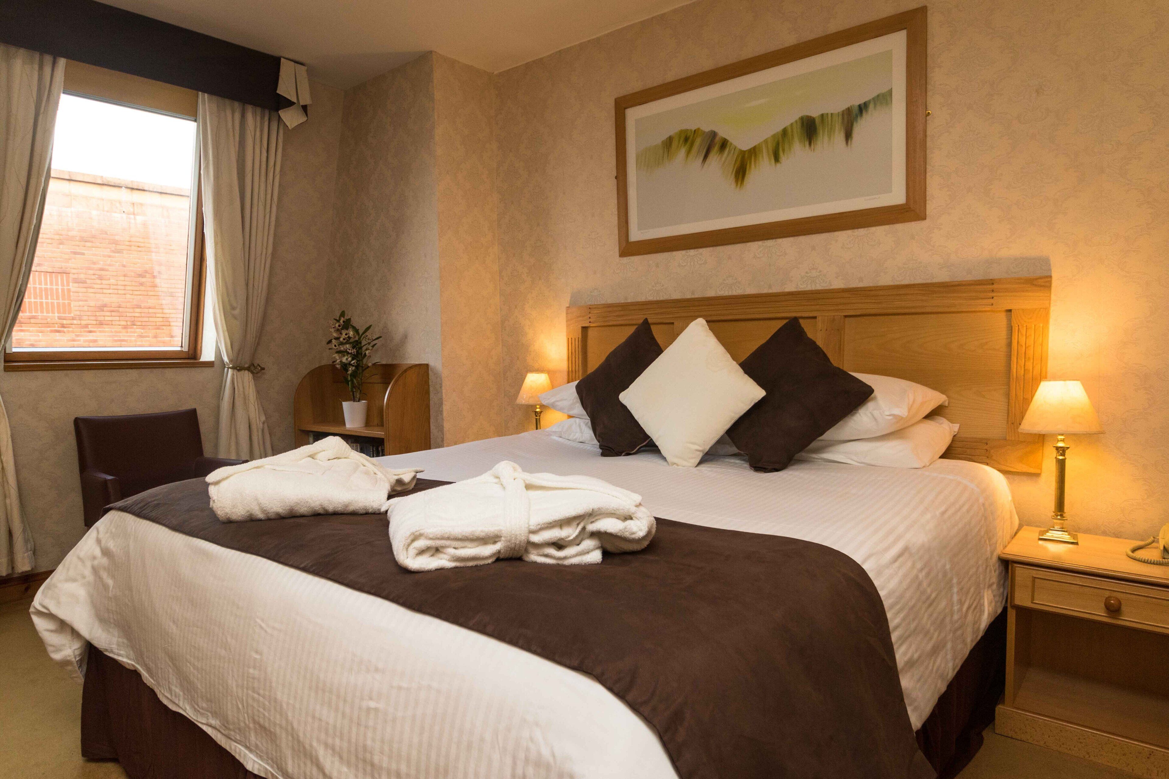 Quarto de hotel com cama grande, duas luminárias de cabeceira, mesa de apoio, uma cadeira e uma janela com cortinas.