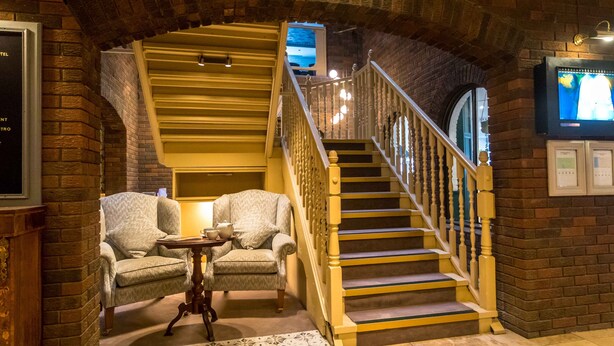 Una escalera con barandilla de madera y pared de ladrillo. Hay dos sillones y una mesita con lámpara.