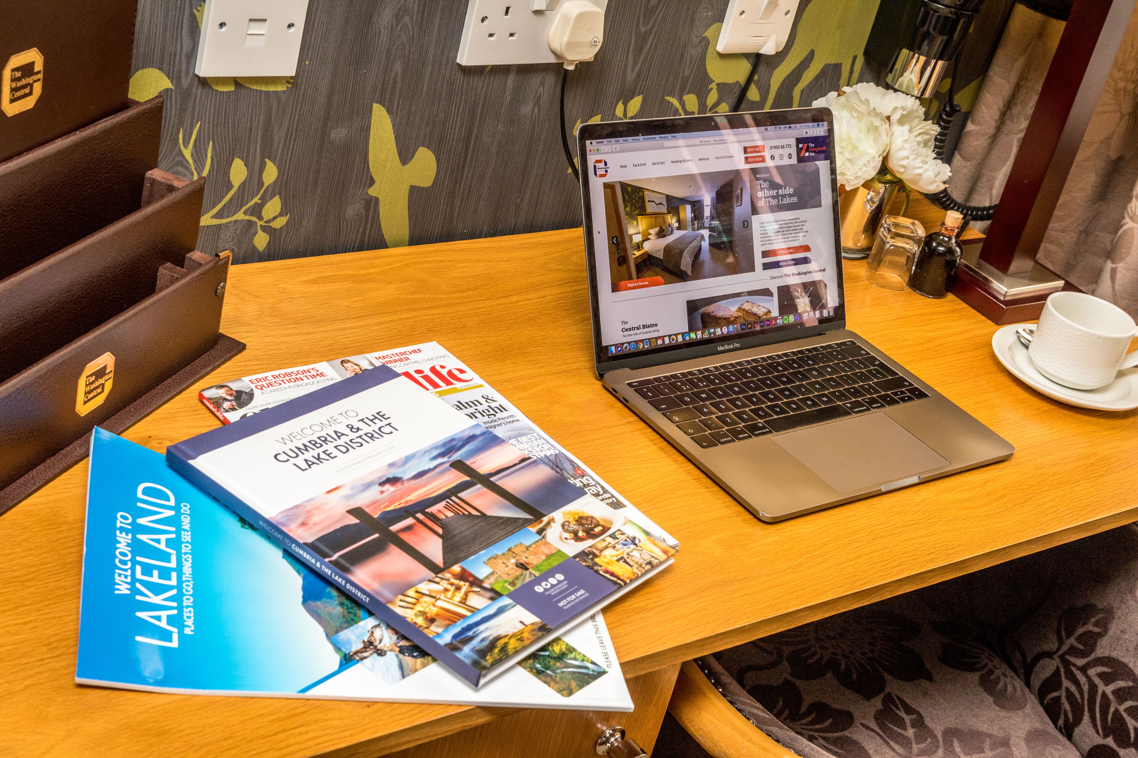 A laptop on a wooden table with a brochure and a cup of coffee.